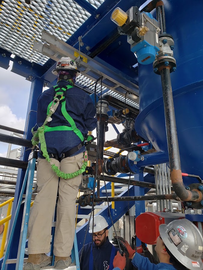 Instrumentation tech in a green fall harness working elevated pipe and valves