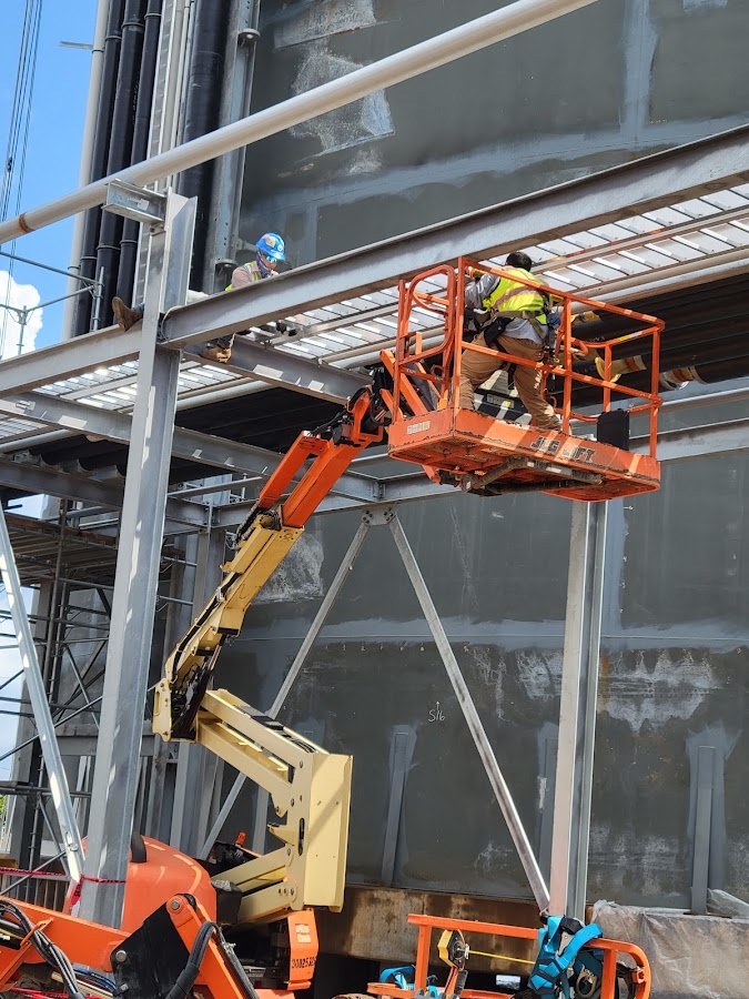 Crew on steel structure with orange boom lift and safety harness in foreground
